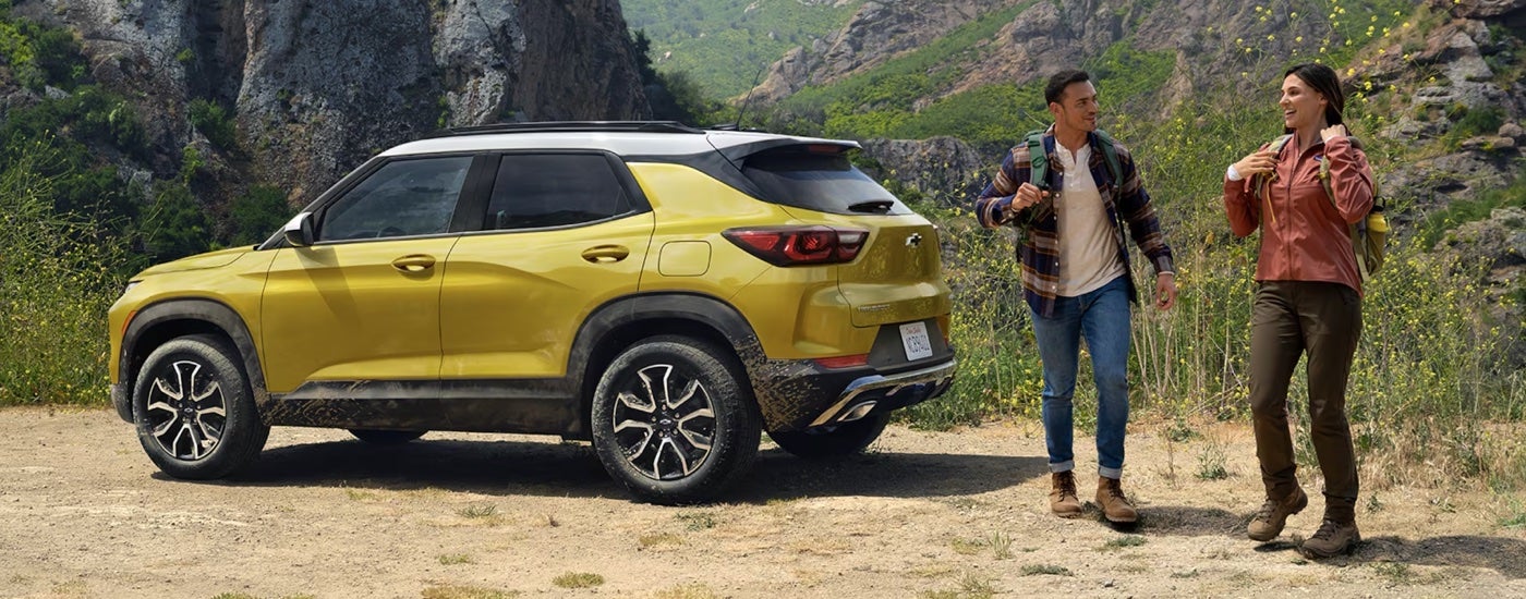 Two hikers walking away from a yellow 2024 Chevy Trailblazer ACTIV parked at a trailhead.